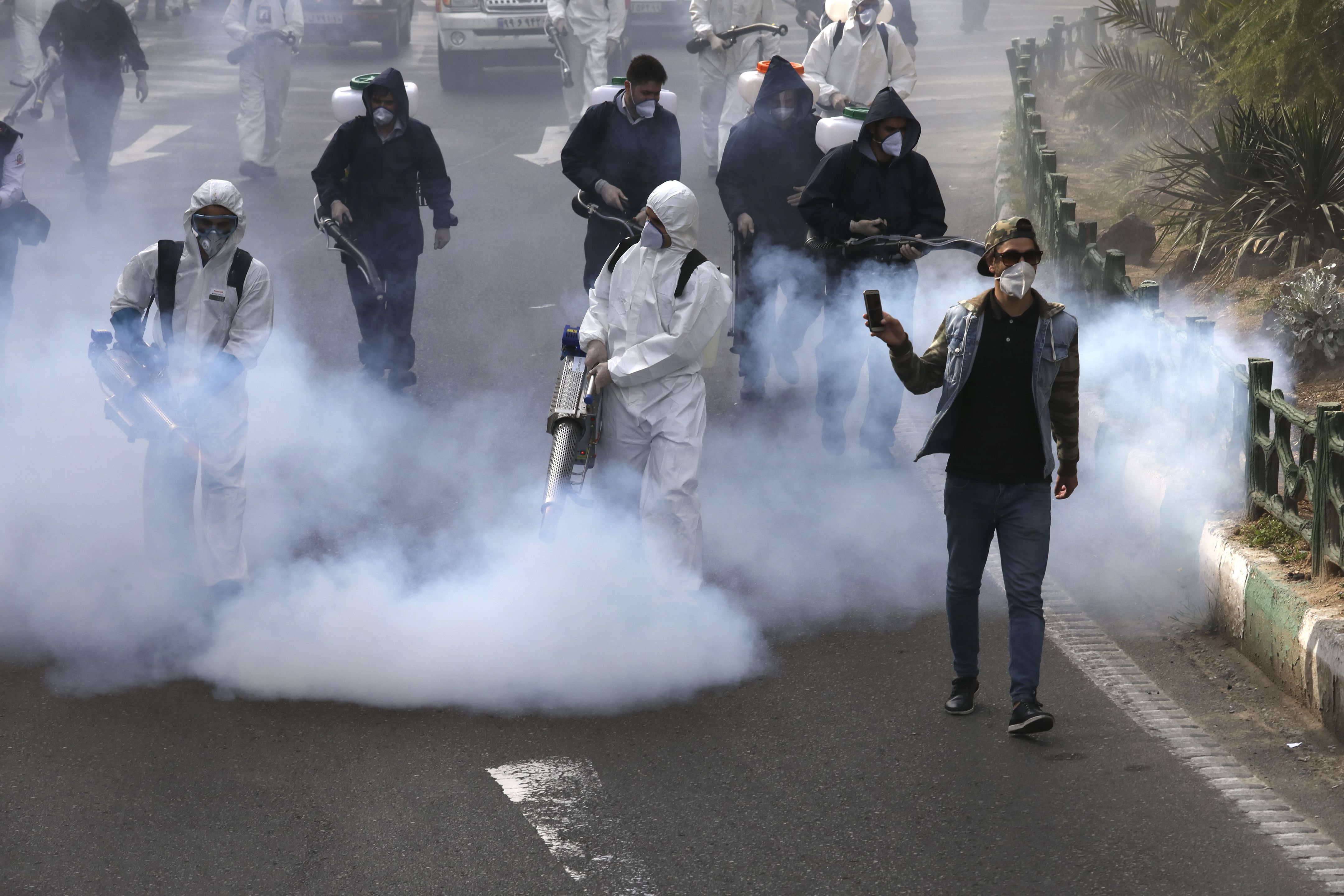 Fotografía del 13 de marzo de 2020 de bomberos desinfectando una plaza mientras un hombre graba video en Teherán, Irán. (AP Foto/Vahid Salemi)