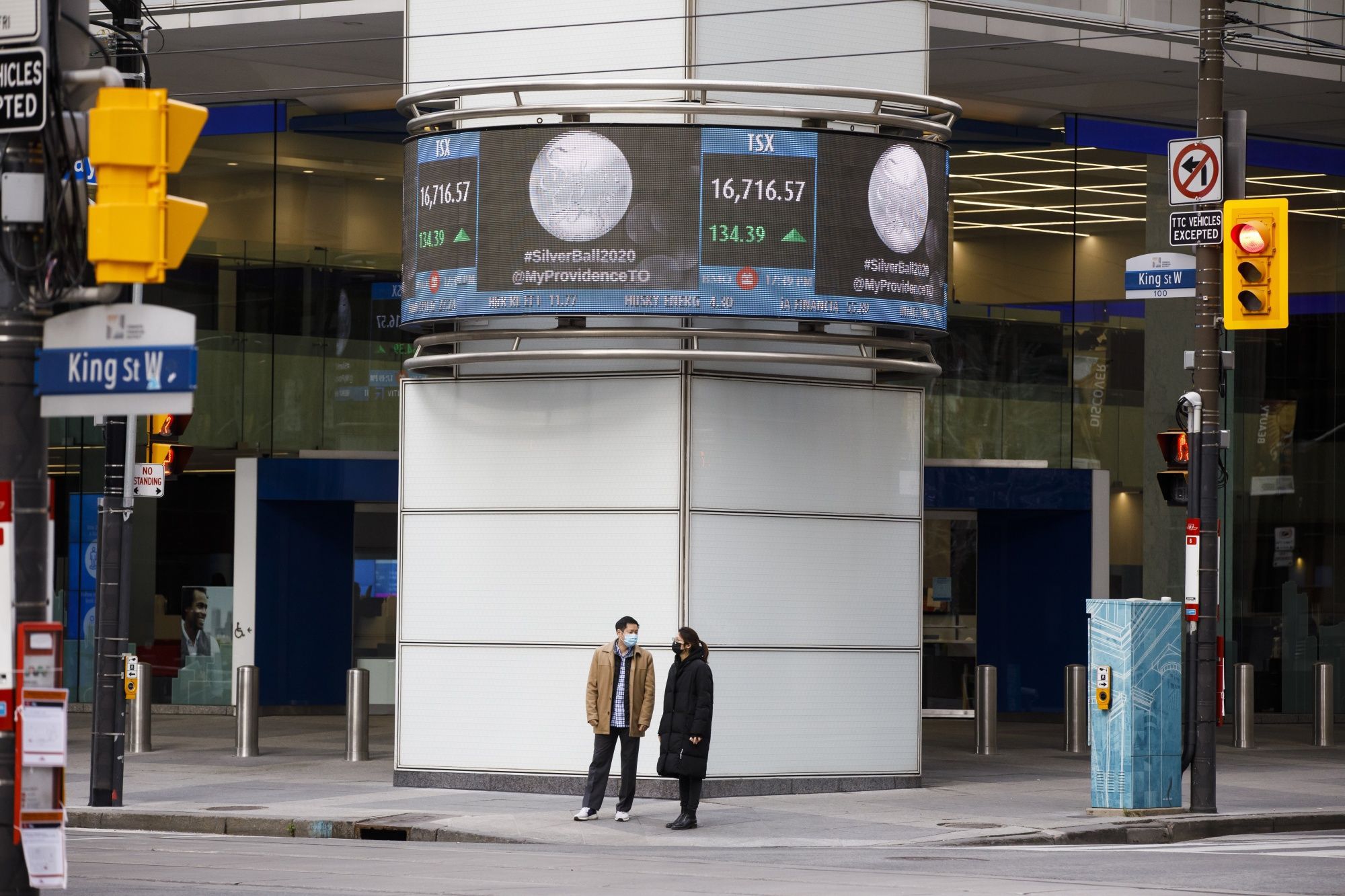 Pedestrians wearing protective masks stand underneath a ticker for the Toronto Stock Exchange in Toronto, Ontario, Canada, on Friday, Nov. 13, 2020. The city of Toronto has said it's forcing casinos, meeting centers, indoor bars and restaurants to stay closed into December as Covid-19 cases continue to rise throughout the city and province.